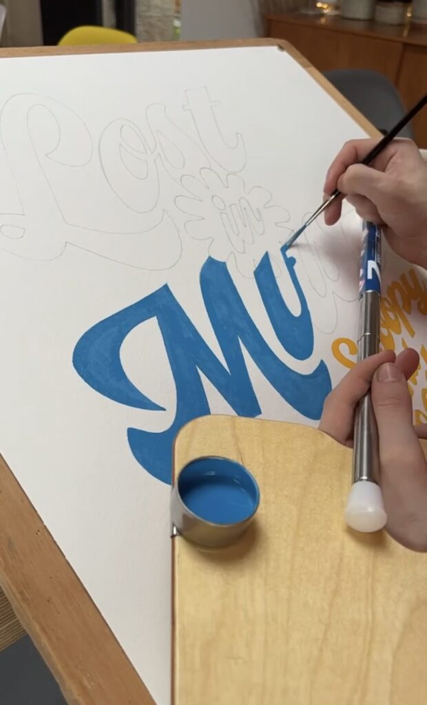 Blue script lettering being painted over a pencil sketch