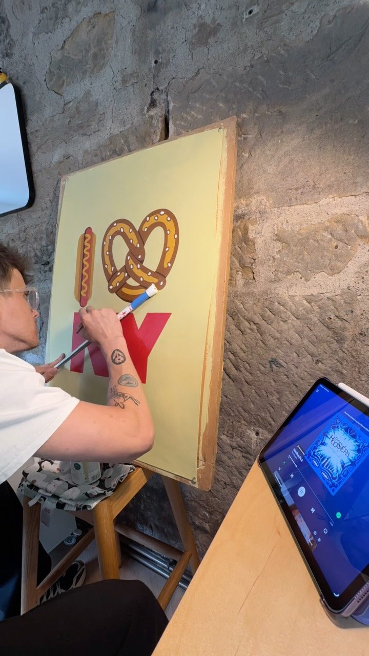 Torin signwriting an I love NY board with a mahl stick at an easel